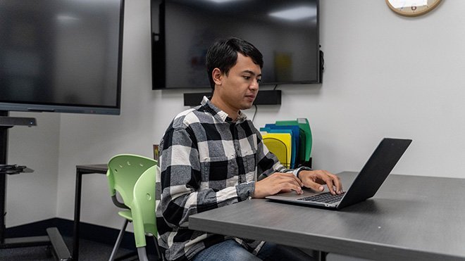 Kyle Villella 鈥�23 sits at a desk and looks at his laptop in the Education Department's model classroom